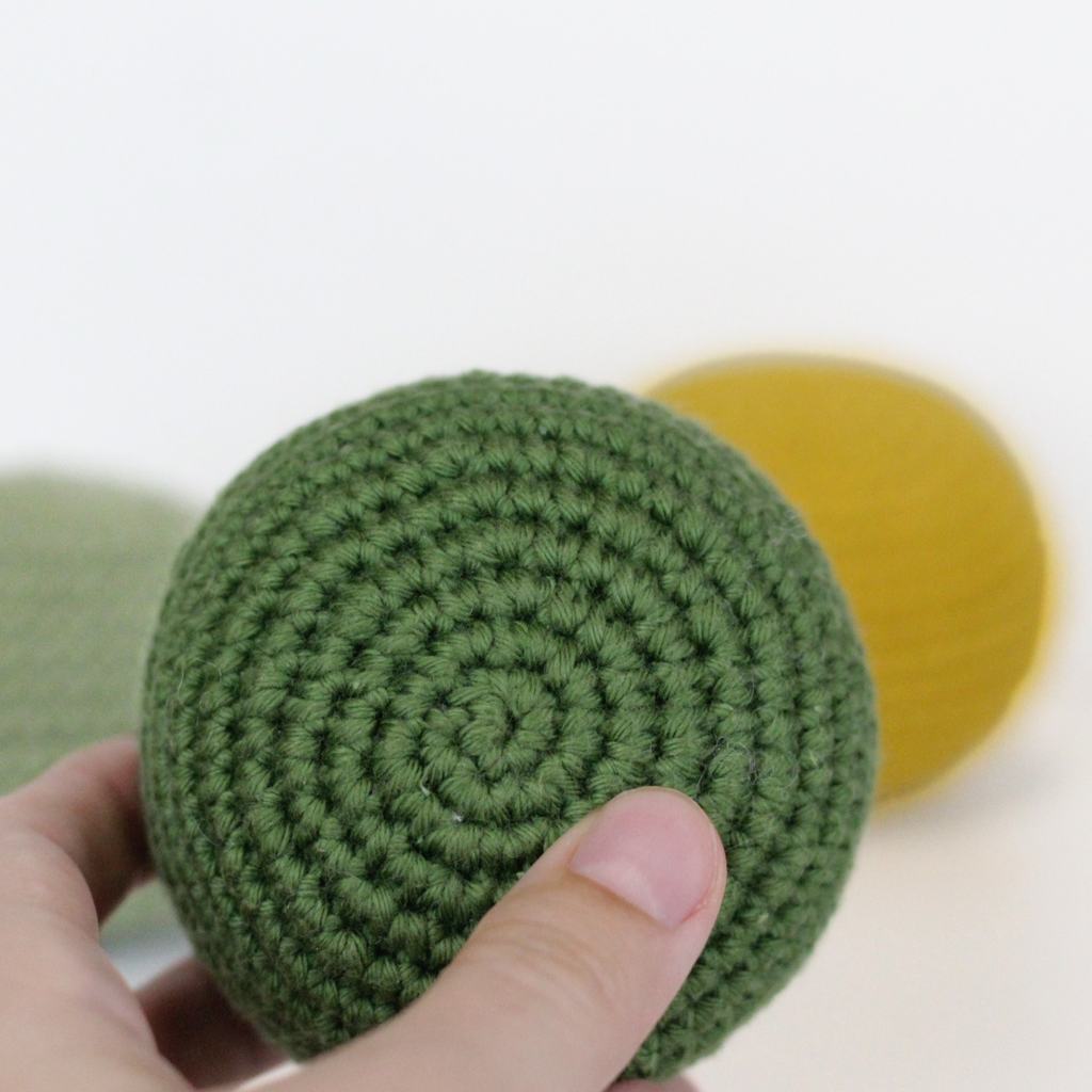 Forest Green crocheted ball held by a hand with a blurred yellow and moss green balls  on the background. Handmade crochet dog toys from Lana Royals Pet Wear.