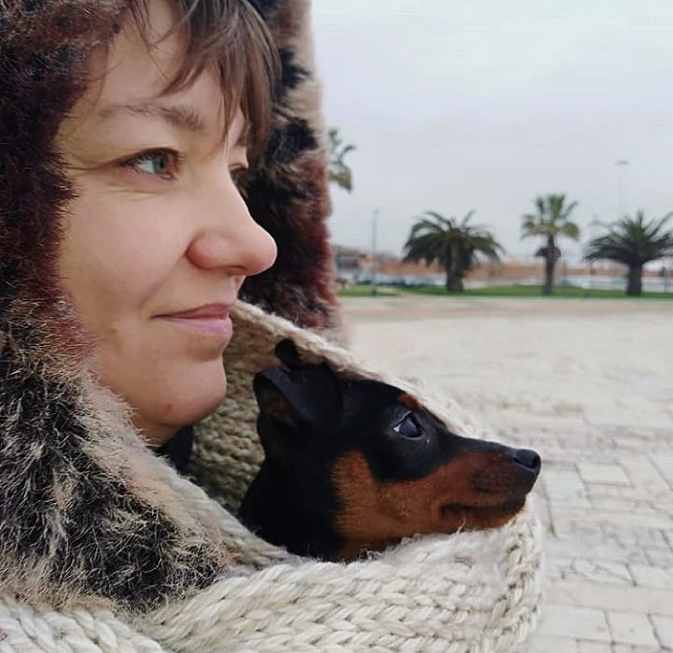 Svitlana from Lana Royals Pet Wear wearing a fur-lined coat with a small dog, Miniature pinscher Rubi peeking out, outdoors on a cloudy day.