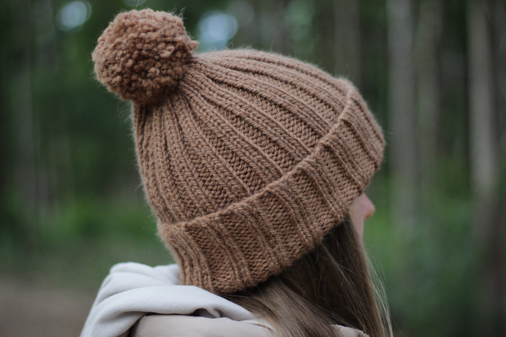 Person wearing a brown hand-knitted thick alpaca wool beanie with a pom-pom in a forest setting. Lana Royals Pet Wear Store.