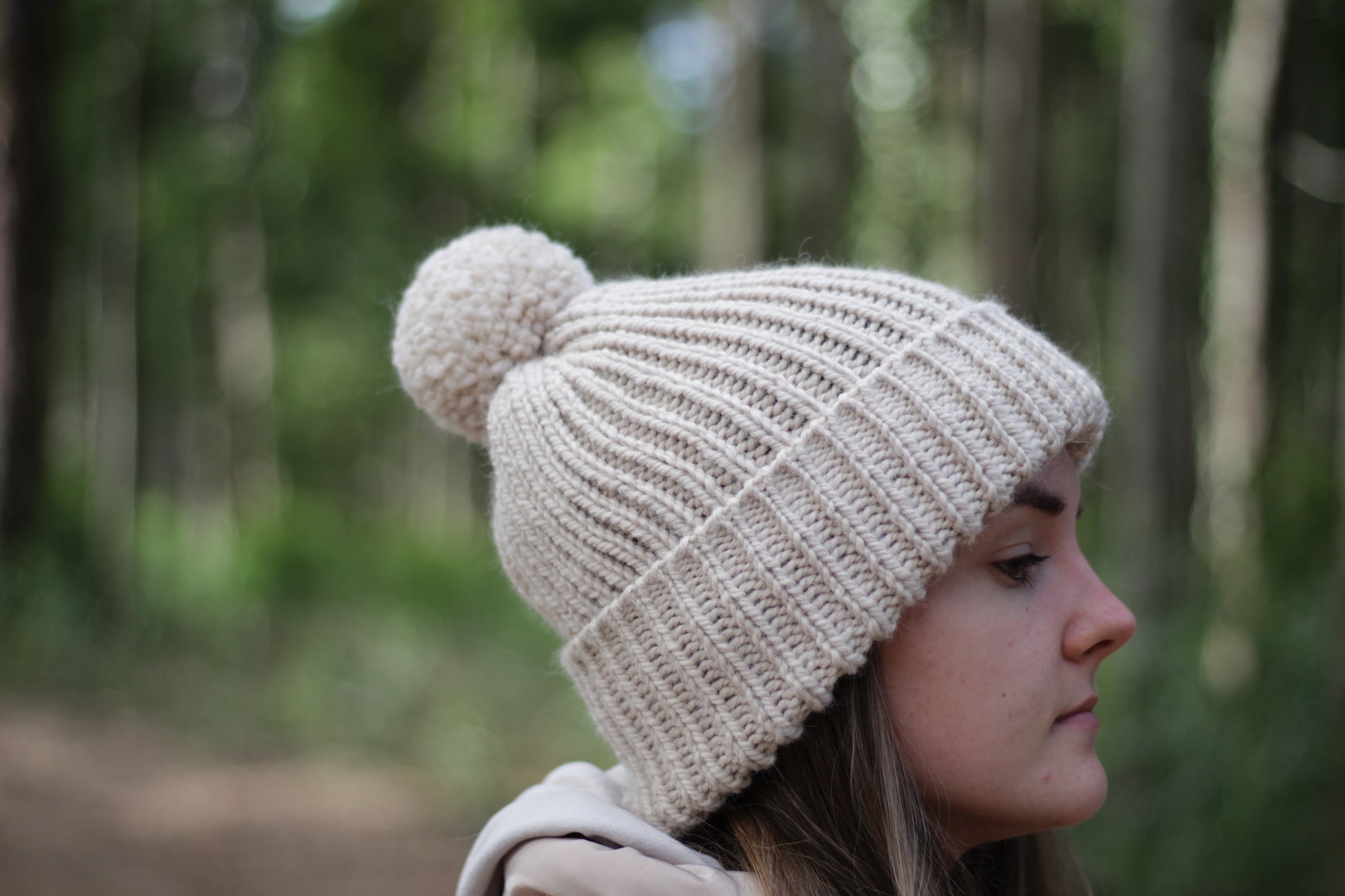 Person wearing a beige hand-knitted alpaca wool hat with a pom-pom in a forest setting. Handmade Lana Royals Pet Wear Store.