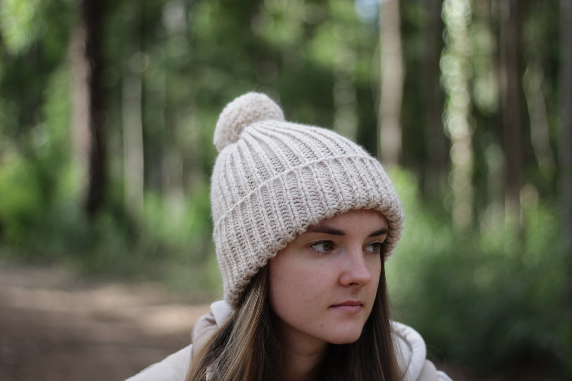 Woman wearing a beige alpaca wool knit hat with pom-pom in a forest setting. Handmade by Lana Royals Pet Wear Store.